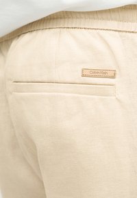 Κοντινή λήψη πίσω τσέπης μπεζ λινών παντελονιών με ελαστική μέση και μικρή ετικέτα Calvin Klein πάνω από την τσέπη.