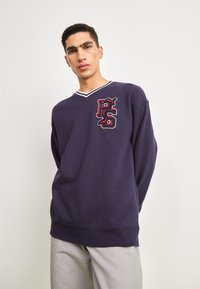 Jūras spēku džemperis ar V veida kakla izgriezumu, kam pievienota sarkana un balta joslu apdare un daudzkrāsains grafiskais logos kreisajā krūtī. Mīksta auduma tekstūra.