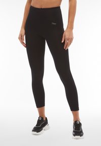 Leggings neri a vita alta realizzati in un tessuto elasticizzato, dotati di una tasca laterale e un sottile dettaglio di branding. Abbinati a sneakers bianche e nere.