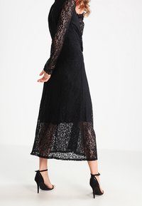 Kvinna i en lång svart spetsklänning med genomskinliga ärmar och nederkant, matchad med svarta högklackade ankle-strap skor, stående på vit golv.