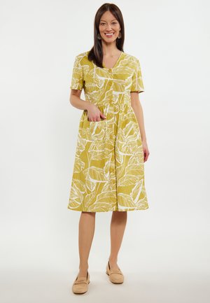 Gelbes Kleid mit weißen Blattmustern, kurzen Ärmeln, V-Ausschnitt, Knopfleiste und seitlichen Taschen. Kombiniert mit hellbeigen Slippern.