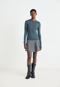 Haut turquoise à manches longues avec une coupe ajustée, associé à une jupe plissée grise. Des bottes noires montantes avec des accents de boucles complètent la tenue.