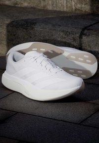 Ζευγάρι λευκά αθλητικά παπούτσια Adidas με ανοιχτό μπεζ σόλες, ένα παπούτσι όρθιο και το άλλο ξαπλωμένο στο πλάι σε πέτρινα σκαλιά.
