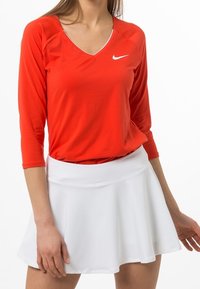 Punainen, pitkähihaisten urheilu paita, jossa on V-kaula-aukko ja valkoinen Nike-logo, yhdistetty valkoiseen hameeseen, joka on väljästi leikattu ja sileäpintainen.