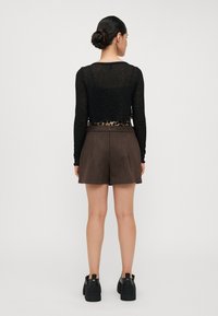 Svart, langermet, gjennomsiktig topp med en leopardmønstret midjebånd, parret med brune, høytlivde shorts og svarte sneakers, sett fra baksiden.