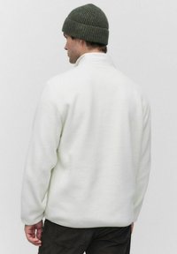 Witte fleece jas met een hoge kraag en een losse pasvorm; heeft lange mouwen en een gestructureerd, zacht oppervlak. G gedragen met een donkergroen muts.