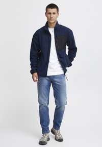 Marine fleece jack met een zwarte ritssluiting, een wit T-shirt eronder, lichtblauwe jeans en grijze wandelschoenen.