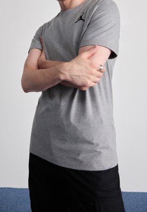 Personne portant un t-shirt Jordan gris, les bras croisés, montrant une bague à la main gauche, sur un fond uni.