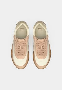 Μπεζ και κρεμ αθλητικά παπούτσια με υφασμάτινο επάνω μέρος, στρογγυλεμένη μύτη, επίπεδα κορδόνια και καφέ λαστιχένια σόλα. Το λογότυπο της μάρκας εμφανίζεται στο εσωτερικό.