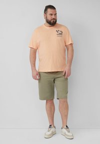 Helles oranges T-Shirt mit einem Grafikdruck eines Palmenbaums, kombiniert mit olivgrünen Shorts und weißen Sneakern mit beigen Akzenten. Glatte Textur überall.