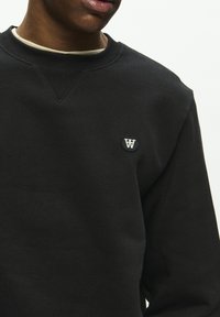 Black sweatshirt featuring a crew neckline, ribbed cuffs, and a small round logo on the chest. Fabric appears soft and textured.