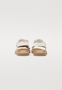 Sandalias blancas con una plantilla suave, correas ajustables y suela de goma marrón. La vista trasera destaca el diseño abierto y el estilo minimalista.