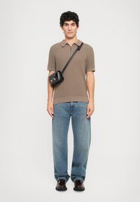 Mann iført brun kortermet genser med glidelås ved kragen, løse blå jeans, svart crossover-veske og mørkebrune sko som står mot en hvit bakgrunn.