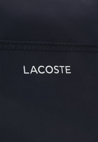 Marinblått tyg med en slät yta som har den vita "LACOSTE"-logotypen. Sömmarna är snygga och jämna längs kanten.