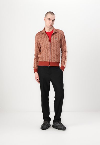 Veste zippée marron à motifs avec une doublure rouge et des accents en rayures blanches, associée à un pantalon noir et des baskets sombres.