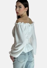 Weiße schulterfreie Bluse mit rüschenförmigen Kanten, strukturiertem Stoff und tailliertem Schnitt. Kombiniert mit hellen Jeans.