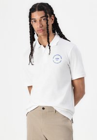 White polo shirt made of cotton with a classic collar, featuring a blue logo print on the left chest, paired with beige pants.