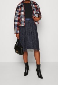 Veste en polaire à carreaux rouge, bleu et blanc ; haut noir ajusté ; jupe plissée marine scintillante ; sac à main structuré noir ; bottines noires.