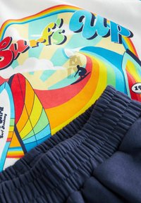 Balta marškinėlių su spalvinga "Surf's Up" grafika, kurioje pavaizduotas banglentininkas, saulė ir vaivorykštiniai bangos. Priekyje - tamsiai navais elastingi šortai.