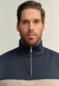Half-zip sweatshirt in navy and beige. Made of soft fabric with a structured collar and a silver zipper. Features a horizontal color block design.