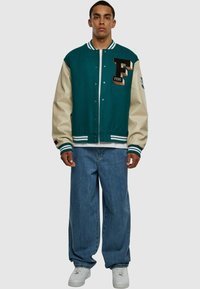 Jachetă letterman în nuanțe de teal și bej, cu embleme, nasturi și manșete cu dungi, purtată cu blugi evazați și pantofi sport albi.