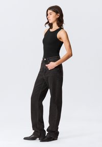 Mulher com top preto sem mangas, jeans largos escuros e sapatos pretos pontiagudos, em perfil, com uma mão no bolso, contra um fundo simples.