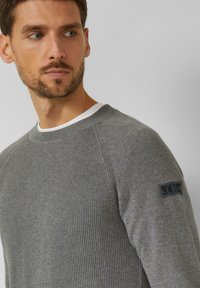 Uomo con capelli castani corti e barba che indossa un maglione grigio lavorato a maglia con una toppa sulla manica sinistra, che guarda verso sinistra su uno sfondo neutro.