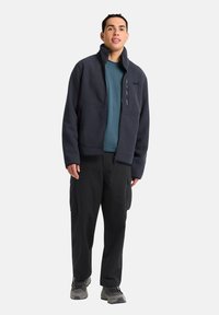 Giacca in pile di colore blu scuro con colletto alto, zip frontale e tasche laterali, indossata sopra una maglietta teal e accoppiata con pantaloni cargo neri e scarpe grigie.
