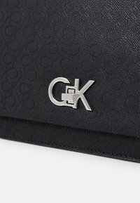 Svart strukturert veske med et mønstrete overflate og en metallisk sølvlogo som detalj, med et elegant, rektangulært design.
