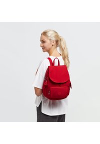 Mochila de tela roja con forma estructurada, tapa redondeada, bolsillo frontal con cremallera y correas ajustables. Presenta un logotipo y un borlón decorativo.