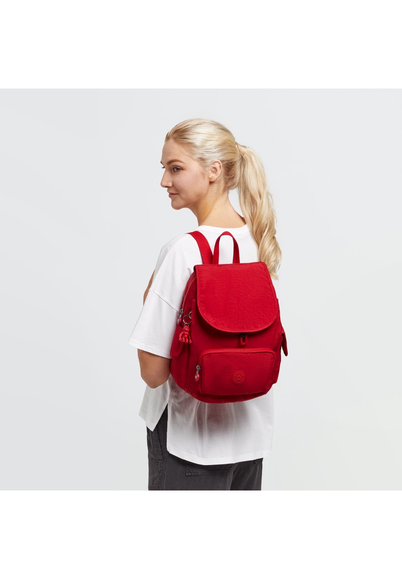 Mochila de tela roja con forma estructurada, tapa redondeada, bolsillo frontal con cremallera y correas ajustables. Presenta un logotipo y un borlón decorativo.