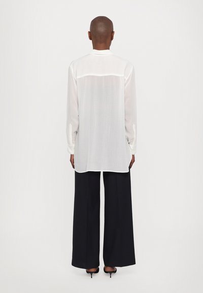 White, long-sleeve shirt with a relaxed fit, featuring a buttoned collar and a sheer back section, paired with navy pants.