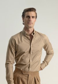 Camisa beige de botones con cuello y mangas largas, combinada con pantalones marrones con pliegues. Tejido suave con un ligero brillo.
