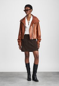 Smeđa kožna skraćena jakna s širokim ovratnikom, bijeli pleteni top s detaljima vezanja, smeđi sued suknja i visoke crne čizme.