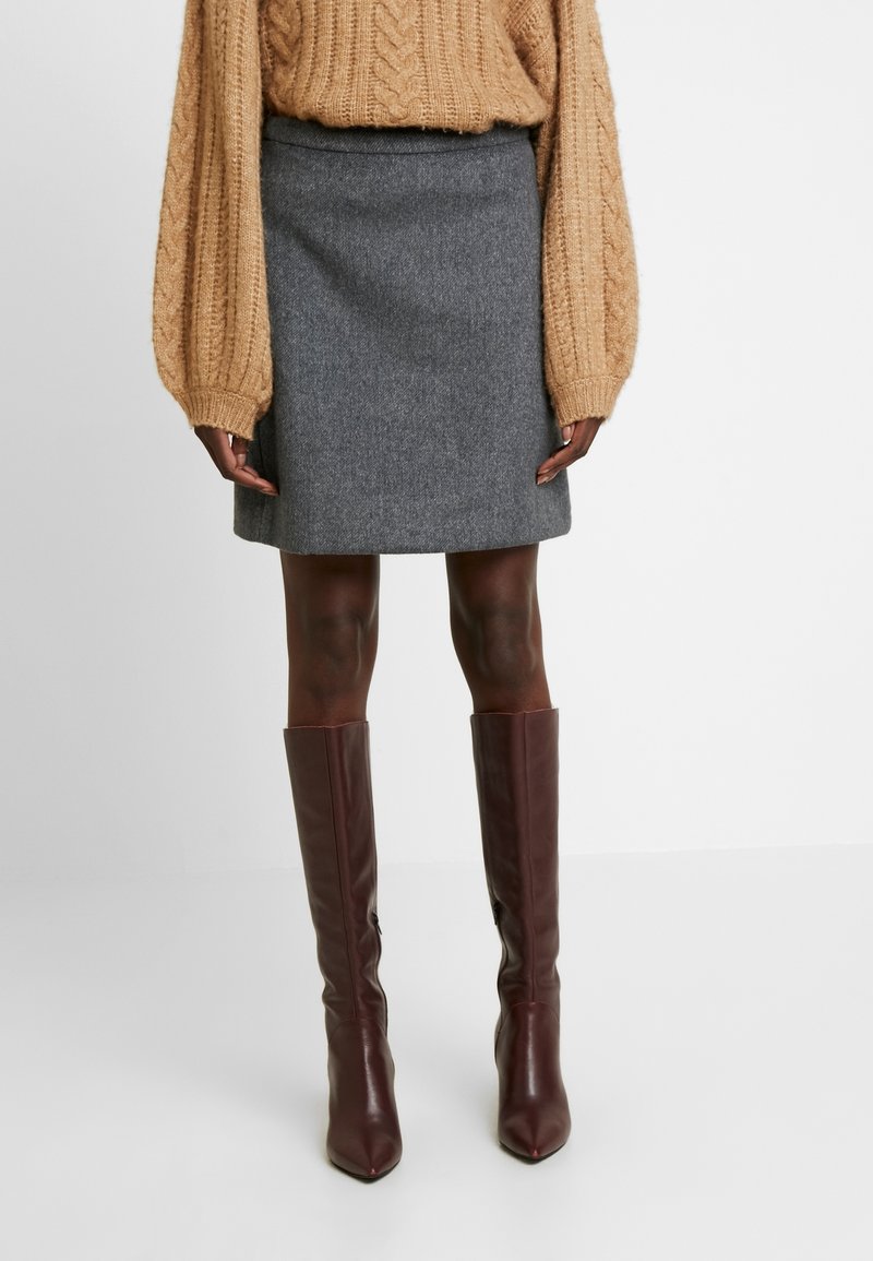 Personne portant un pull en maille torsadée couleur camel, une mini-jupe en laine grise et des bottes en cuir marron foncé montant jusqu'aux genoux, debout sur un fond blanc uni.