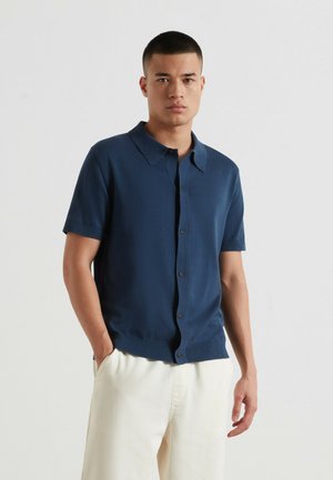 Mann trägt ein marineblaues Kurzarm-Hemden mit Knöpfen und weiße Hose, steht mit einer Hand in der Tasche vor einem schlichten Hintergrund.