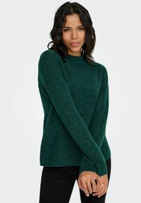 Adīts zaļš džemperis ar augstu apkakli un garām piedurknēm, raksturīgs teksturētai audumam un brīvam piegriezumam. Kombinēts ar melnām pieguļošām biksēm.