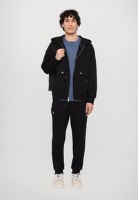 Sudadera negra con cremallera y dos bolsillos frontales, combinada con una camiseta azul y pantalones jogger negros con bolsillos laterales. Zapatillas blancas en los pies.