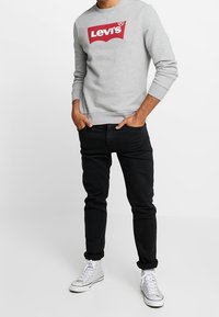Felpa grigia con logo rosso Levi's, abbinata a jeans neri e sneakers bianche. Presenta polsini a costine e un design con scollo rotondo.