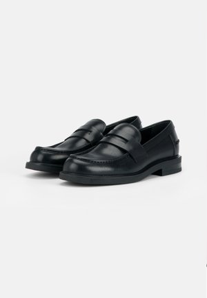 Porą juodų odinių penny loafer batų su žemais kulnais, rodomų ant vienspalvio šviesaus fono.