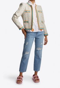 Veste en tweed avec des accents orange et bleu, deux poches avant, associée à une chemise blanche à volants, un jean bleu déchir�é et des sandales roses.