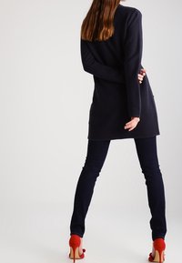 Lång marinblå cardigan med texturerad väv, parat med figurnära marinblå byxor och röda högklackade skor. Bakifrån framhävs ärmlängden och siluetten.