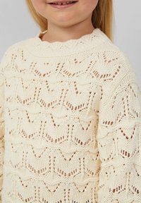 Pull en tricot crème avec un col à volants et un motif ajouré complexe, avec une finition texturée et une coupe ample.