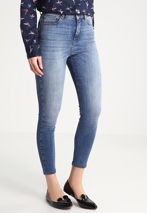 Jeans ajustados de color azul con un lavado desgastado, que cuentan con cinco bolsillos y un diseño de cintura media, combinados con zapatos planos de charol negros.