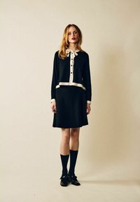 Svart klänning med vit krage och knappar, som har långa ärmar och en figursydd midja. Modellen bär svarta knähöga strumpor och loafers.