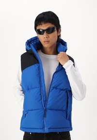 Gilet rembourré bleu avec des panneaux d'épaules noirs, capuche, fermeture éclair avant et poches latérales. Tissu texturé avec une finition brillante. Porté par-dessus une chemise blanche.