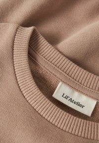 Lichtbruine katoenen trui met een ribgebreide ronde hals en zijnaadstiksels, met een label waarop "Lil'Atelier" in zwarte tekst staat.