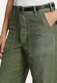 Olijfgroene cargobroek met een relaxte pasvorm, voorzien van zijzakken, zichtbare stiksels en een zwarte riem met een decoratieve zilveren gesp.