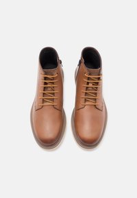 Botas de tobillo de cuero marrón con cordones amarillos contrastantes, textura suave, punta redondeada y detalle de cremallera lateral para facilitar su uso.