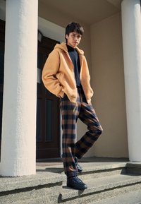 Haut en polaire orange avec zip à l'avant, chemise à manches longues noire, pantalon à carreaux marron et noir, et baskets noires. Debout sur des marches en pierre.